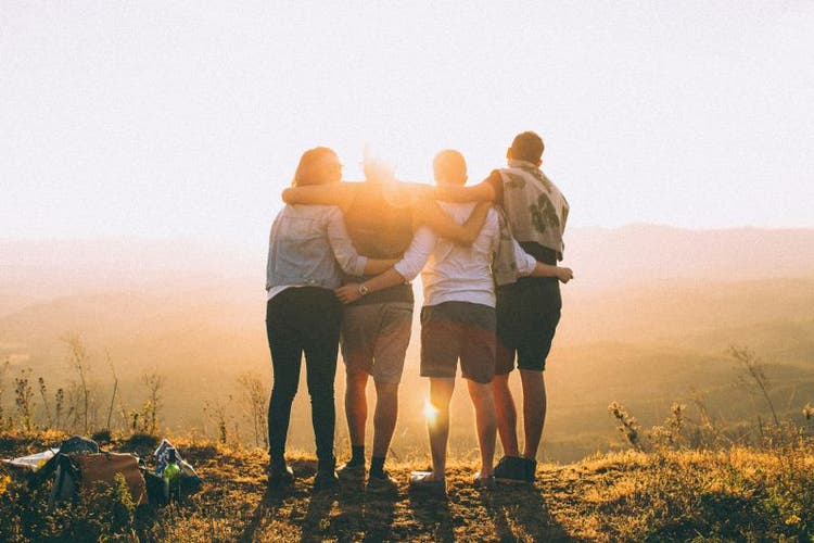 Group of people stand looking at sunset with arms wrapped around each other.