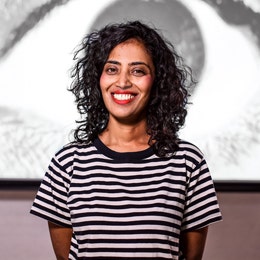 Priya Namana in front of their artwork (partially obscured by Priya's profile, a close up of a human eye in greyscale).
