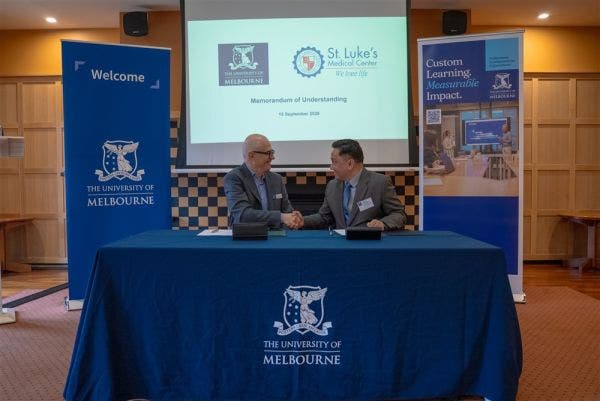 Two men in suits shake hands across a table after signing a Memorandum of Understanding, with a projector screen showing the University of Melbourne and St. Luke's Medical Center logos behind them.