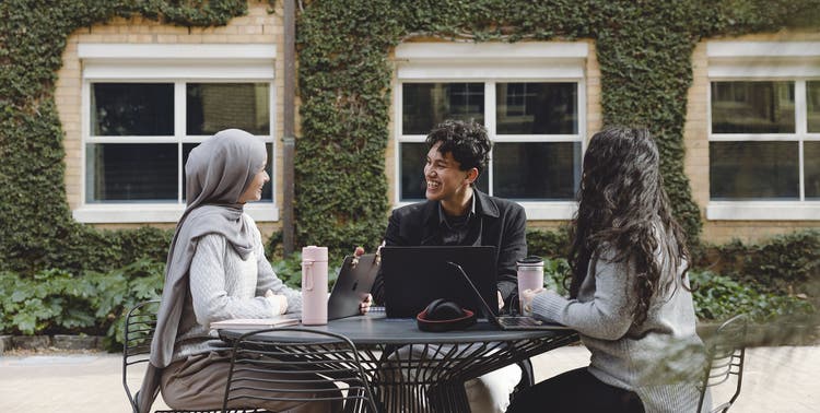 3 students sit at a round table outside, laptops open talking to each other. In the background is a building covered in ivy.
