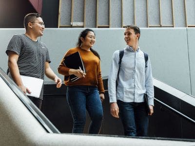 Three students walk down stairs inside a campus building, talking to each other.