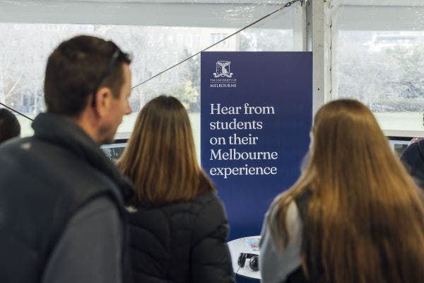 3 people stand in front of a sign that reads Hear from students on their Melbourne experience