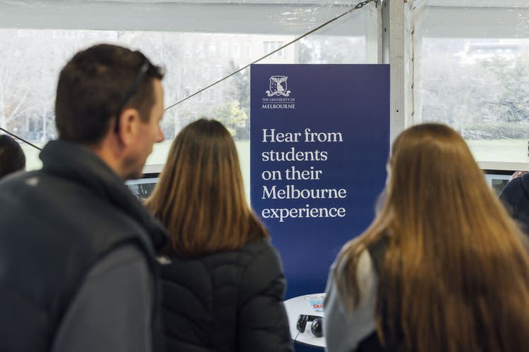 3 people stand in front of a sign that reads Hear from students on their Melbourne experience