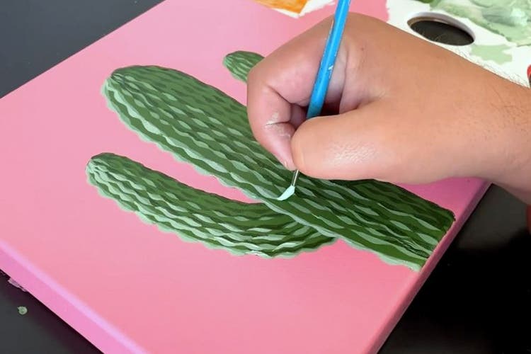 Photo of someone's hand painting a green cactus on a pink canvas.