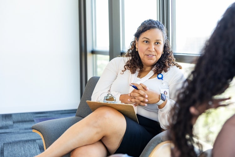 A female social worker sits on a couch holding a clipboard and talking to a young woman.
