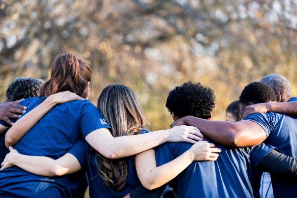 A group of young people, photographed from behind, stand next to each other with their arms wrapped around each others' shoulders.