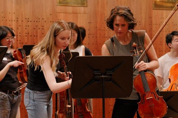 A student and orchestra player stand each holding a violin while pointing and reading a piece of music.