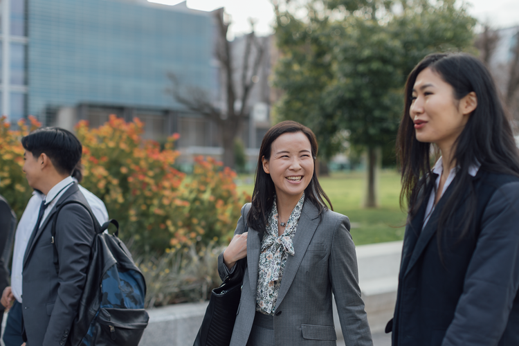 Two students walk and talk smiling on campus, one holds a handbag over shoulder.