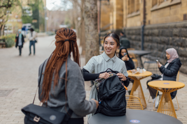Two students pack their back backs, talking and smiling at a table on campus.