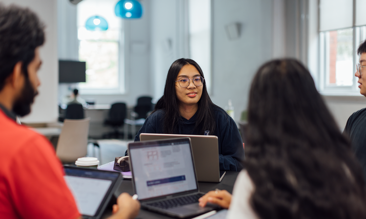 Four students sit together with laptops at a desk indoors.