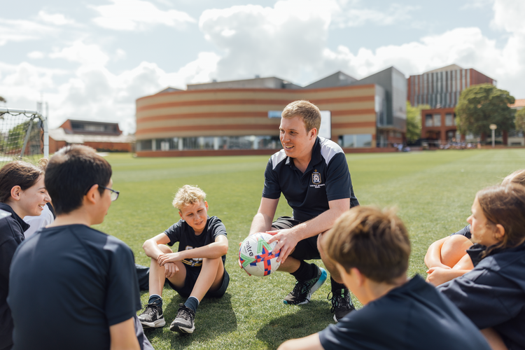 Teacher sits in front of group of students while holding a soccer ball outdoors on a school oval.