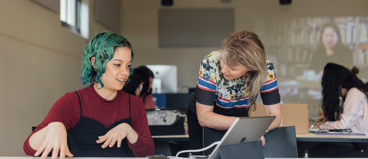 One person sits at a desk with a laptop and talks to another who is standing.