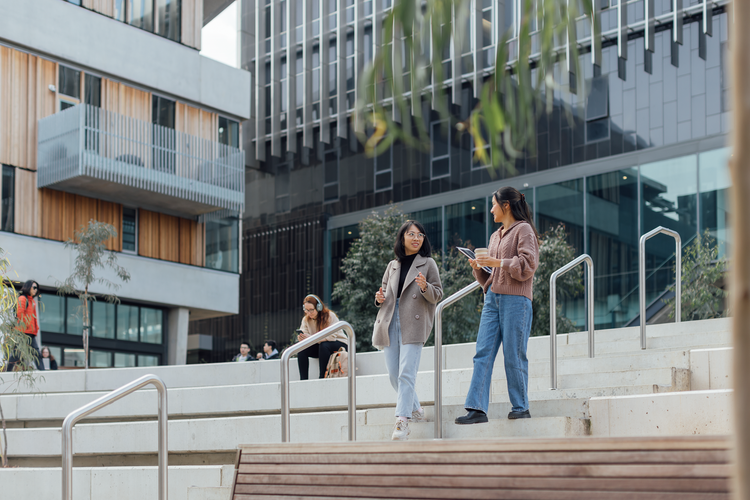 Two students talk smiling on campus walking down stairs and holding study belongings.