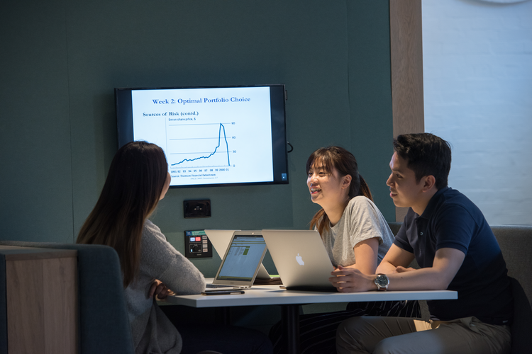 Three students sit together with laptops at a desk in front of large screen.