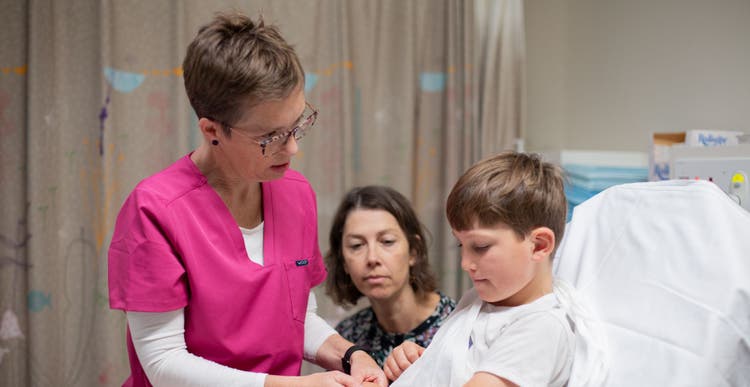 Nurse wearing pink scrubs tends to a young boy on a hospital bed with bandaged arm.