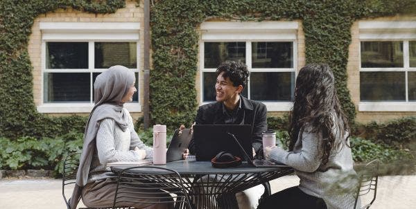 3 students sit at a round table outside, laptops open talking to each other. In the background is a building covered in ivy.