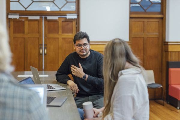 Two people sit talking indoors at a desk with a laptop.