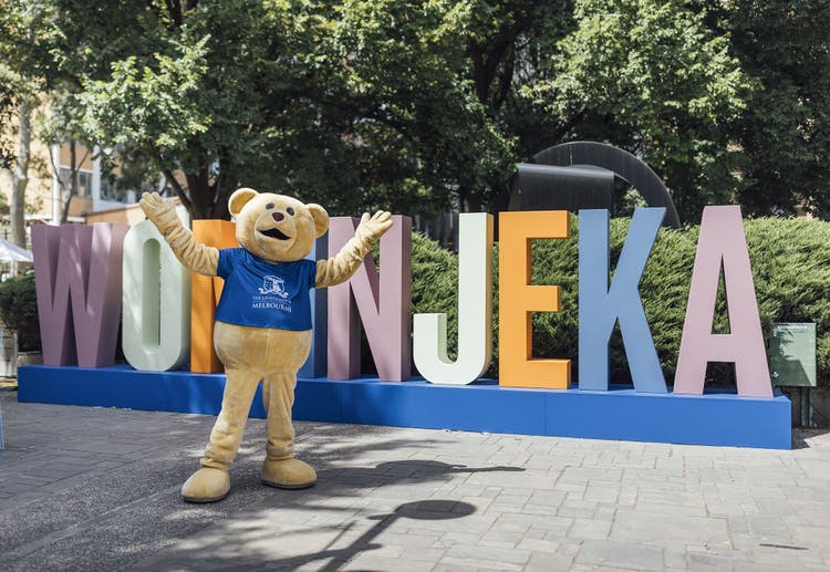 University mascot Barry the bear stands with open arms infront of a sign that reads Wominjeka