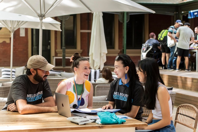 4 people sit at a table laughing with a laptop and clipboard in front of them. Two of the people are wearing Melbourne Orientation tshirts