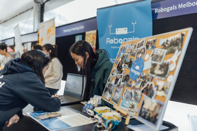 Two students look at a laptop at a Robogals Melbourne display. In the foreground is a pinboard with photos of club activities on it.