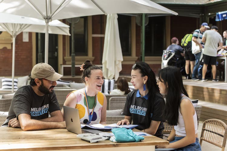 4 people sit at a table laughing with a laptop and clipboard in front of them. Two of the people are wearing Melbourne Orientation tshirts
